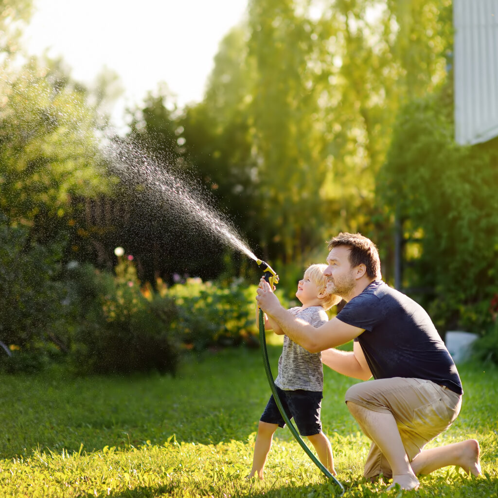 Funny little boy with his father playing with garden hose in sunny backyard