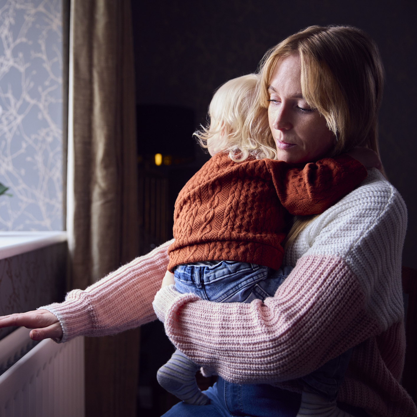 Mother With Son Trying To Keep Warm By Radiator At Home During Cost Of Living Energy Crisis