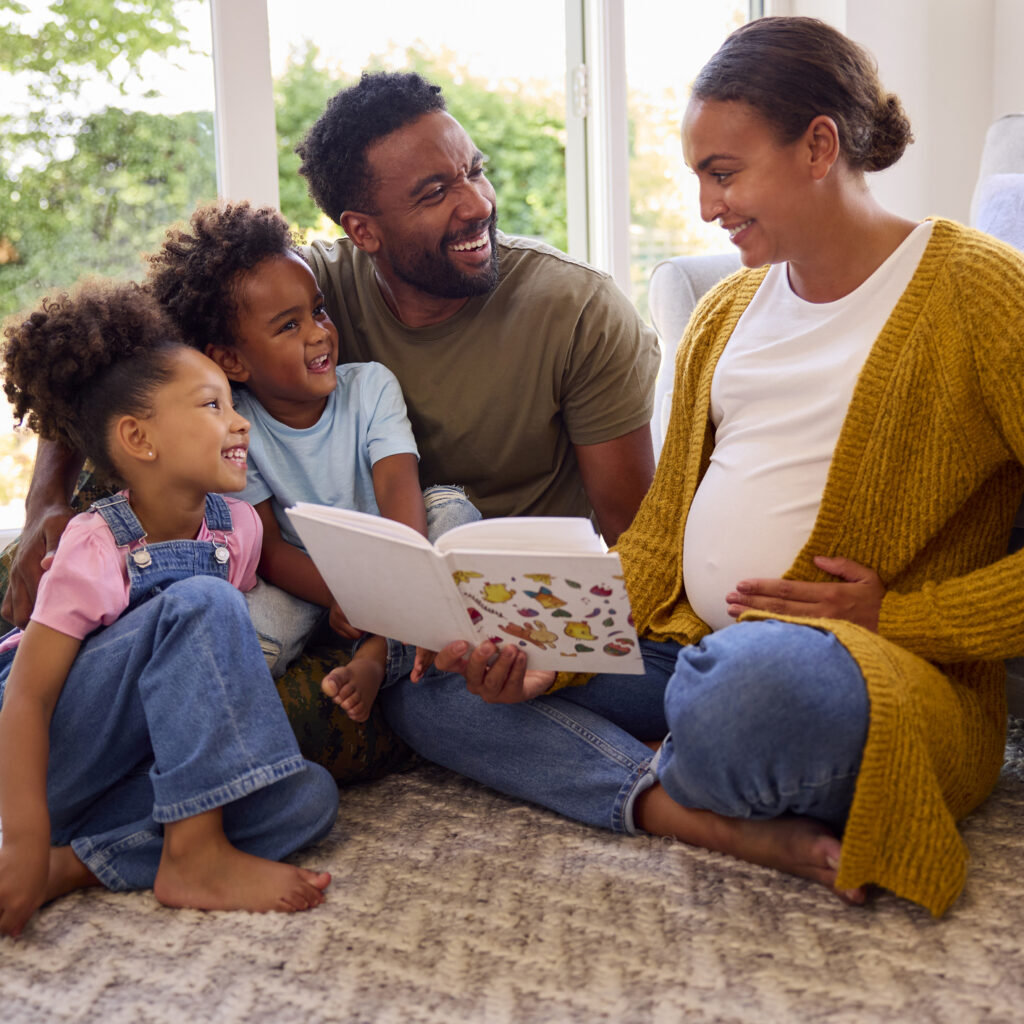 Army Family With Pregnant Mother Sitting On Floor In Lounge At Home Reading Book Together