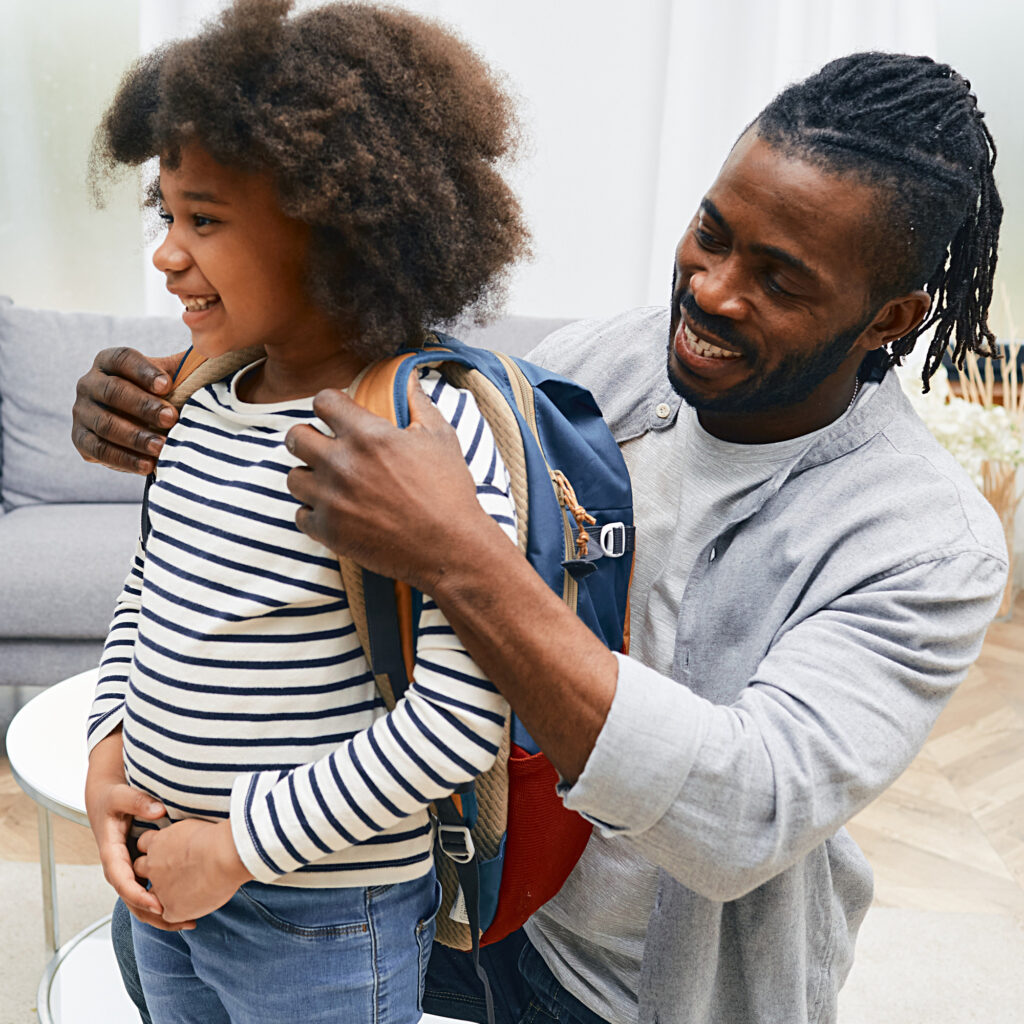 Happy_African_American_Dad_helps_daughter_getting_ready_for_school_and_puts_a_school_bag_on_her_back