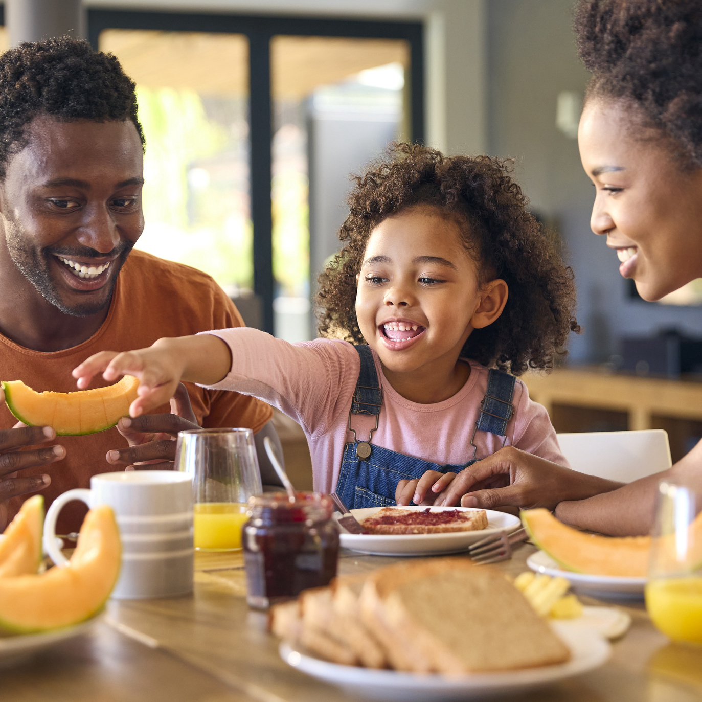 Family Shot With Parents And Daughter At Home Having Breakfast Spreading Jam On Bread At Table