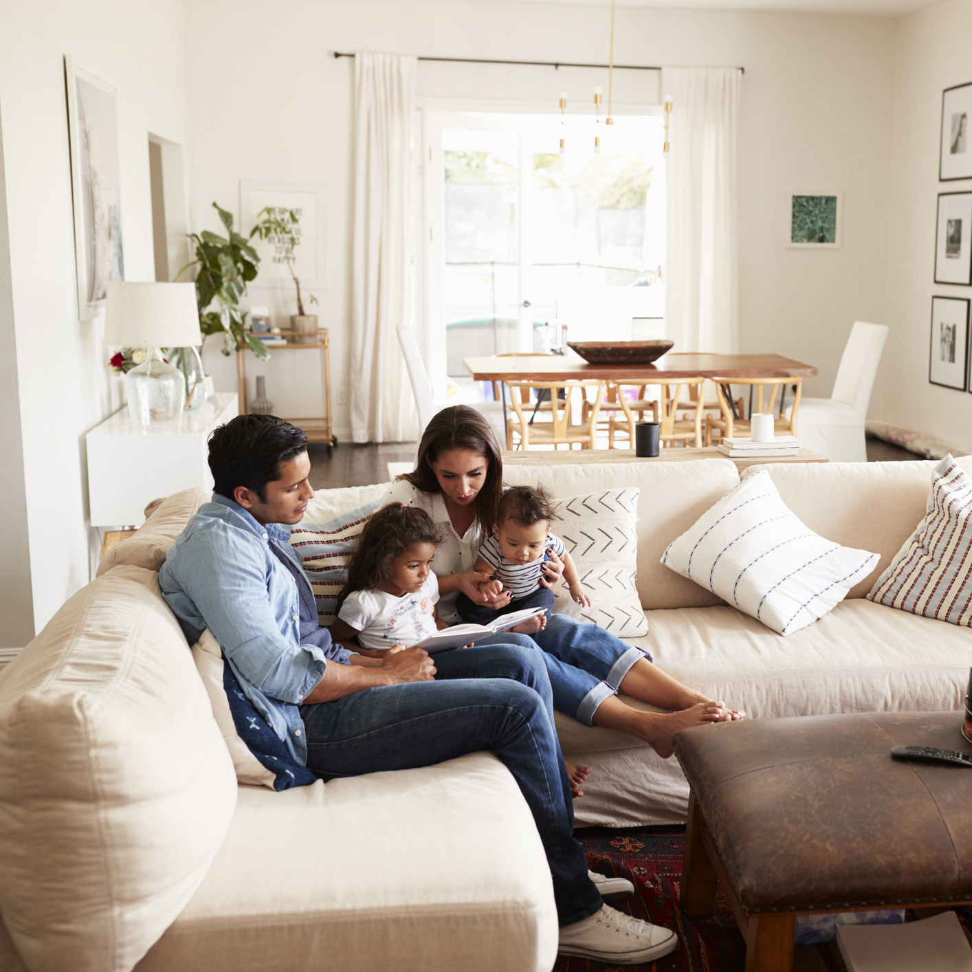 Young Hispanic family sitting on sofa reading a book together in the living room, seen from doorway