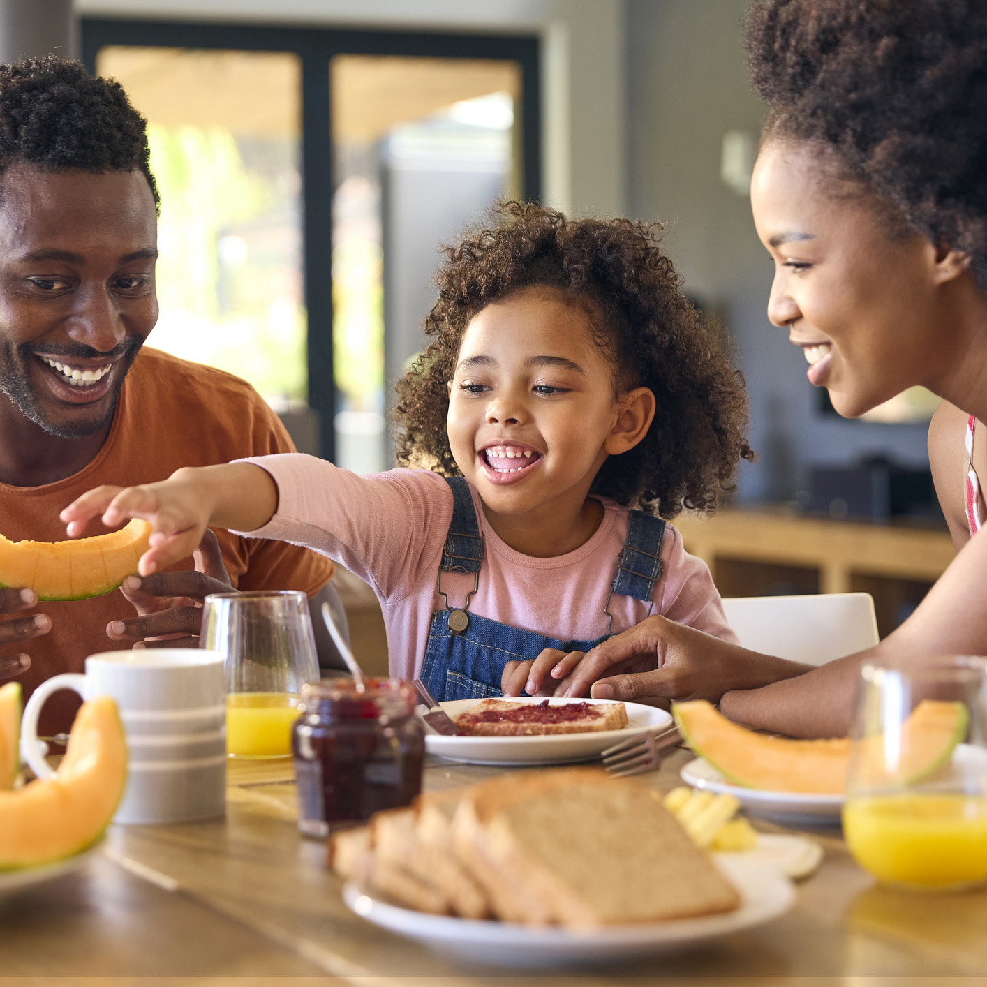 Family Shot With Parents And Daughter At Home Having Breakfast Spreading Jam On Bread At Table