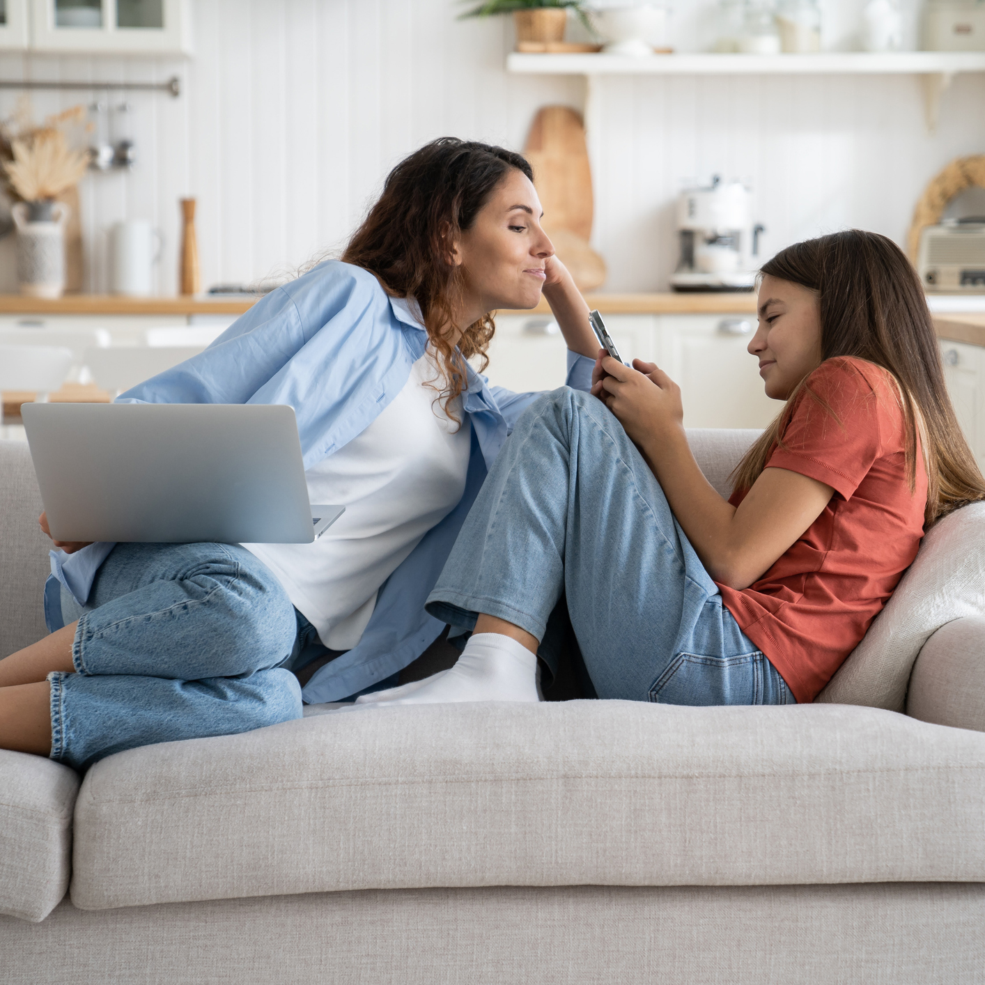 Young smiling woman sitting on sofa with laptop spying on child teen girl using smartphone