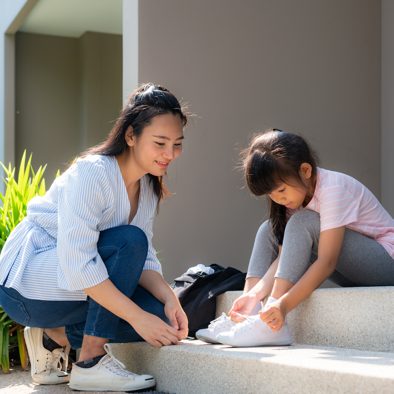 Asian mother help her daughter primary students in uniform to wearing their shoelace of shoes infront of home in the morning school routine for day in the life getting ready for school.